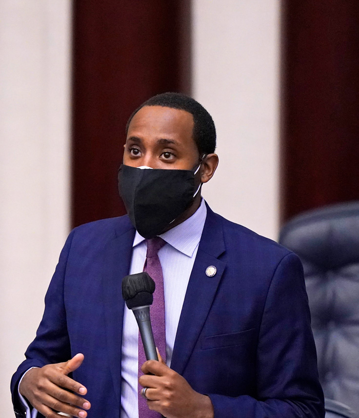 man speaking into microphone, wearing suit and tie, and a face mask