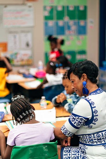 a woman sitting at a child's desk speaking with the child