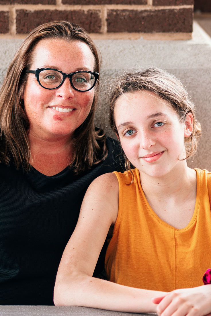 a mom in black, left, and a teen in yellow, right, sitting next to each other in front of brick wall