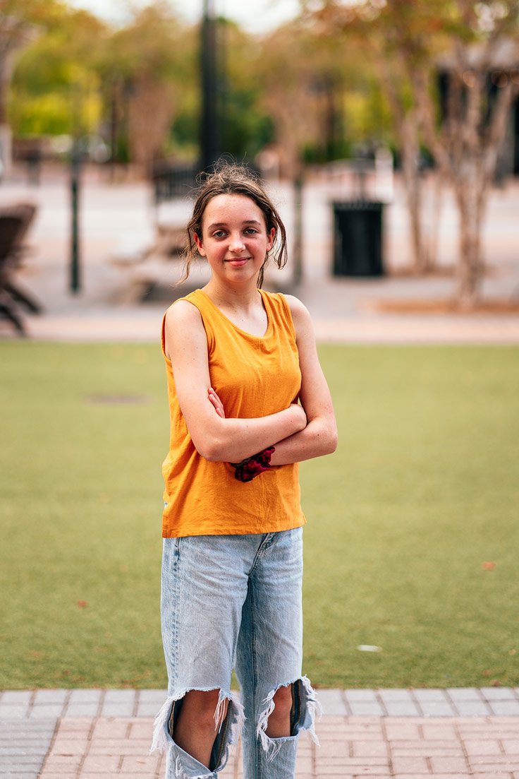 a blonde, blue-eyed  teen in yellow and ripped jeans stands with her arms crossed