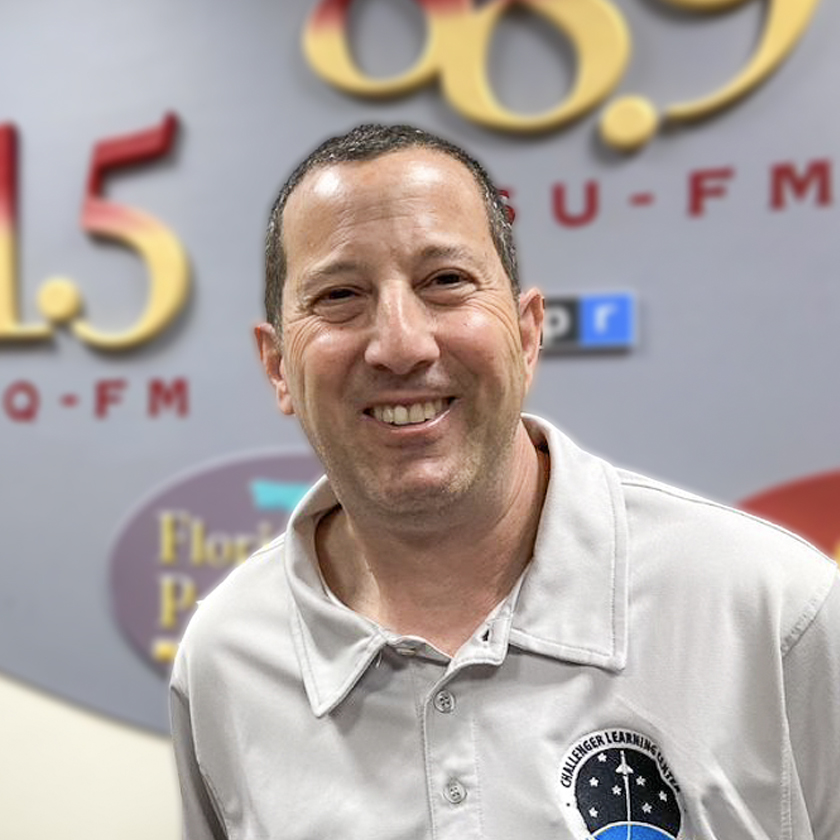 A smiling man stands in front of a  grey background with garnet and gold letters and numbers.