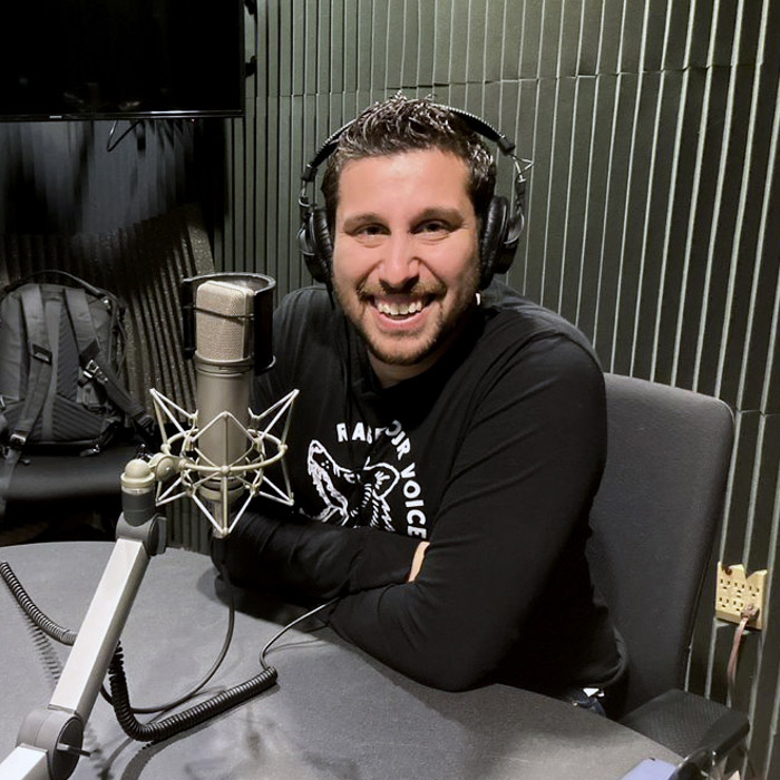 A man dressed in black sits smiling in front of a silver microphone.