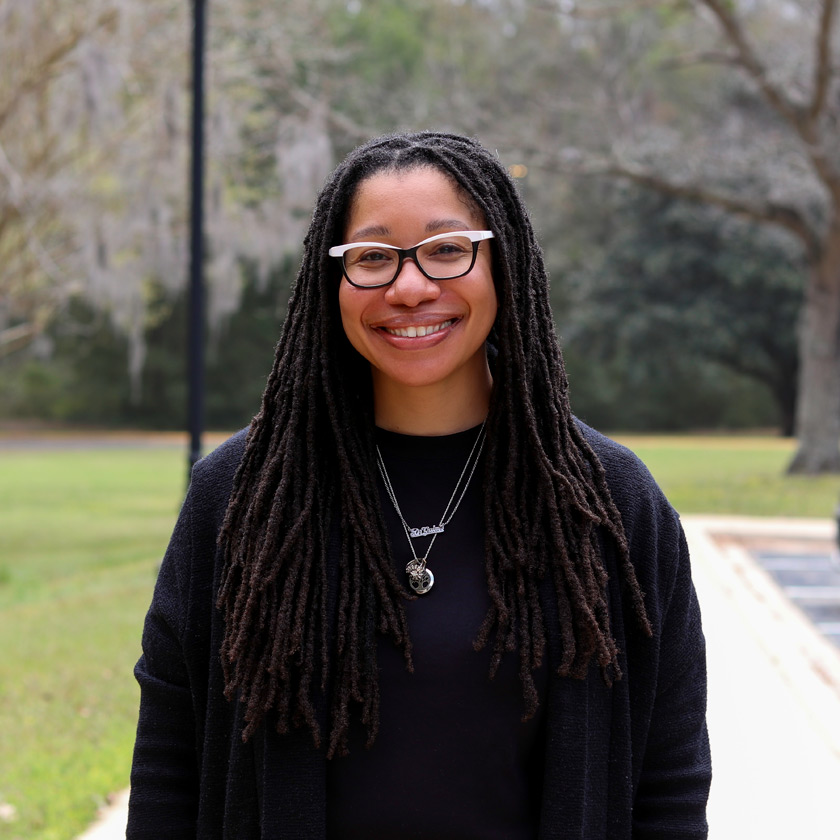 A smiling woman dressed in black stands in front of shrubbery.