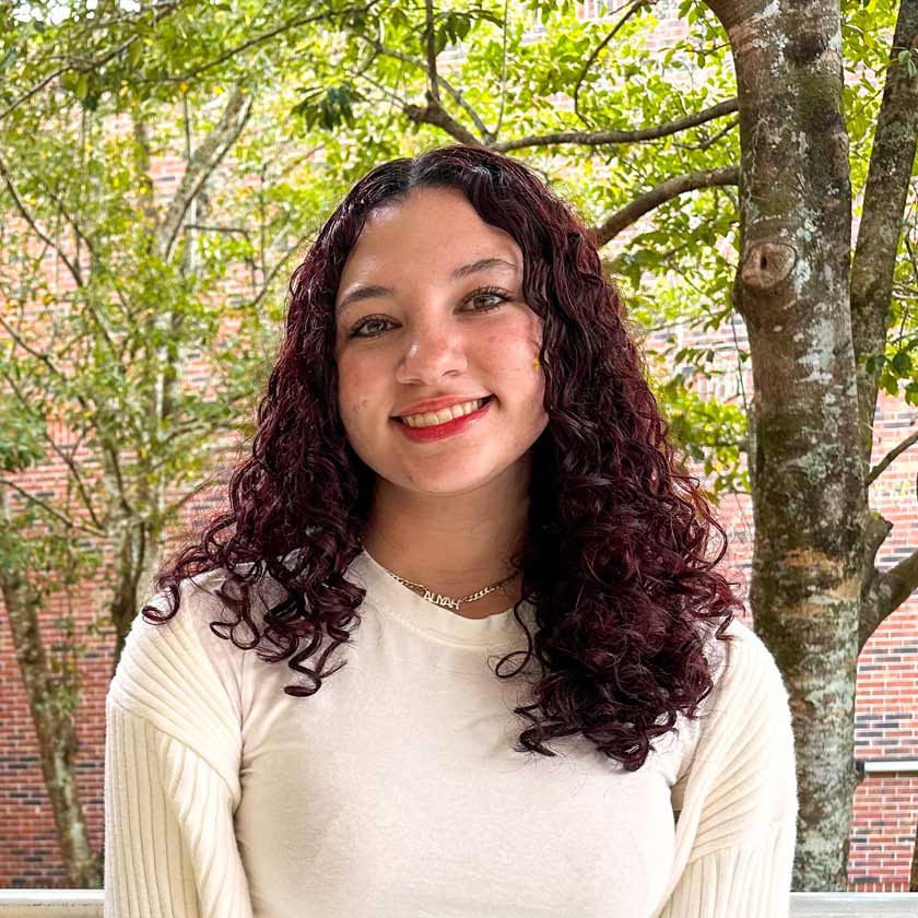 A smiling woman stands in front of a tree and a red brick wall.