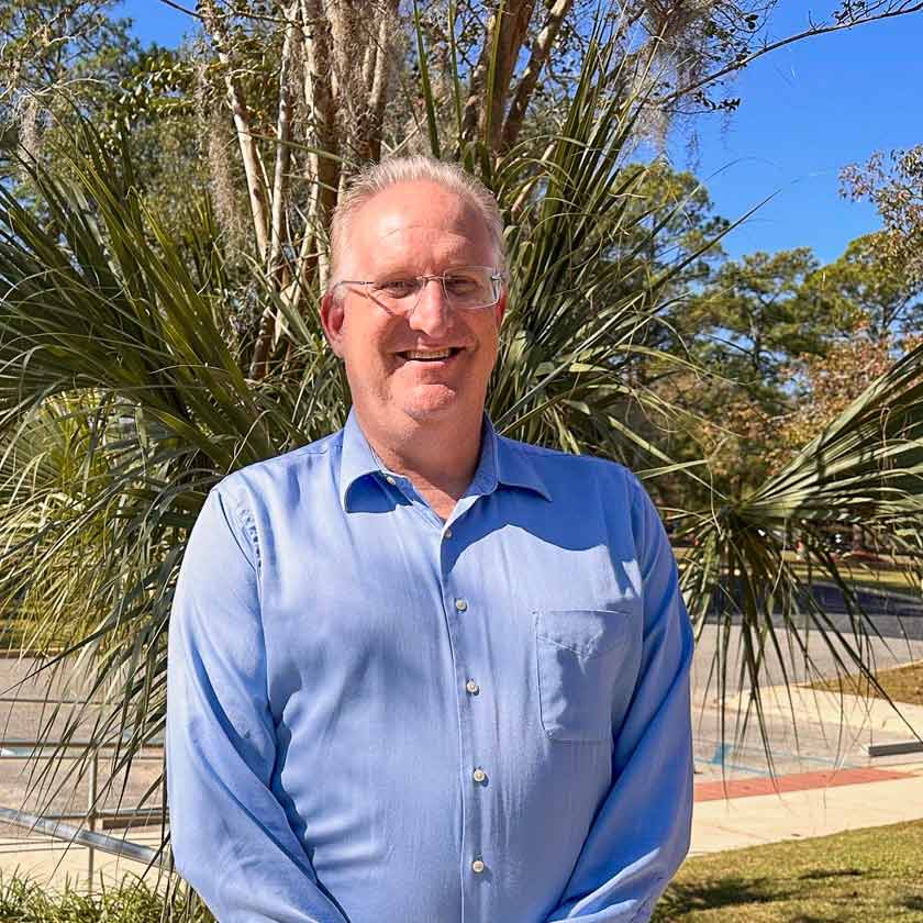 A smiling man stands in front of a bush.