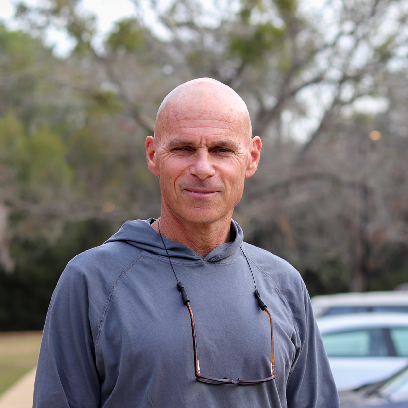 A man in a grey hoodie stands in front of shrubbery.