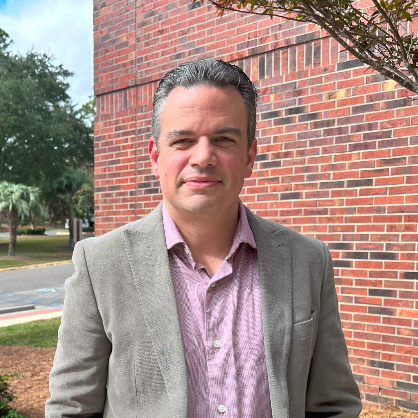 A smiling man stands in front of a red brick wall.