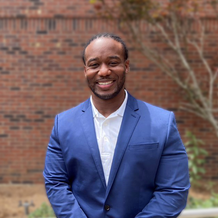 A smiling man in a blue suit stands in front of a brick wall.