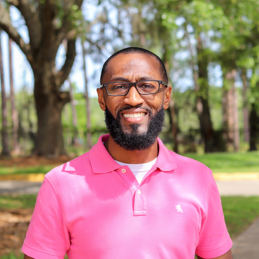 A smiling man in a pink shirt stands outside in front of shrubbery.