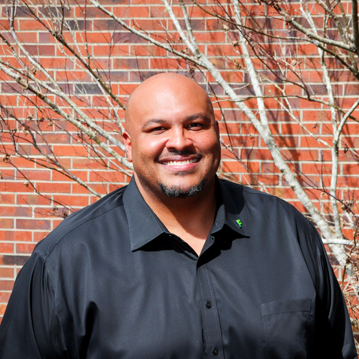 A smiling man in a dark shirt stands in front of a red brick wall.