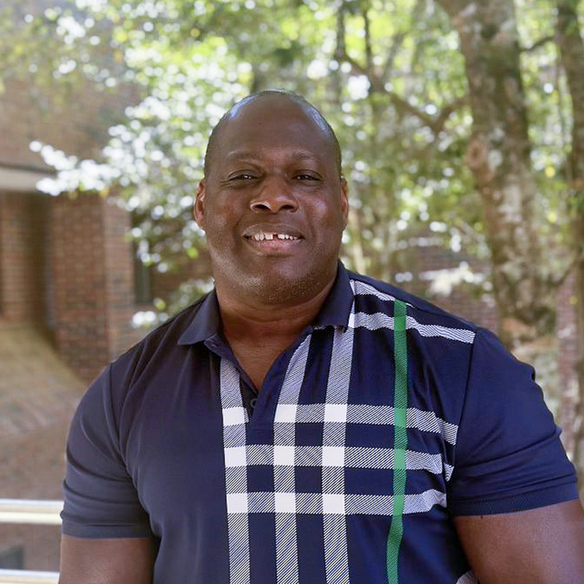 A smiling man in a blue shirt stands in front of a brick wall and a tree.