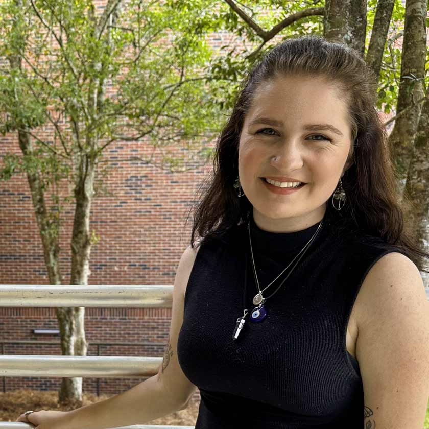 A smiling woman stands in front of shrubbery and a red-brick wall.