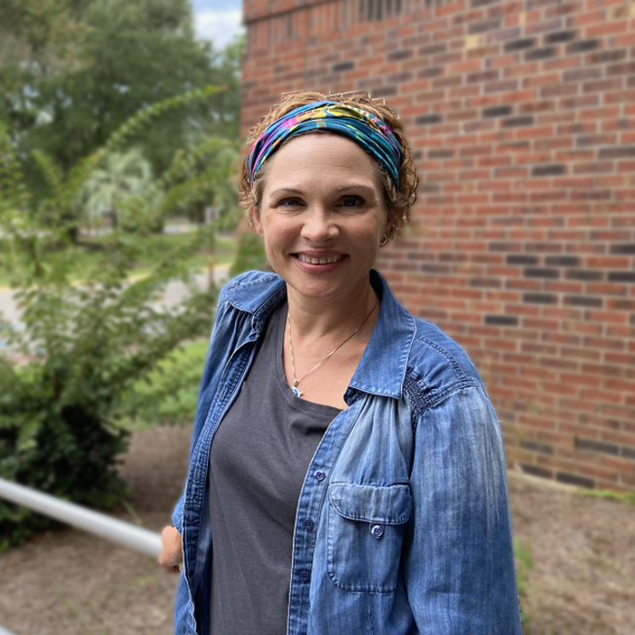 A smiling woman in a jean jacket stands outside.