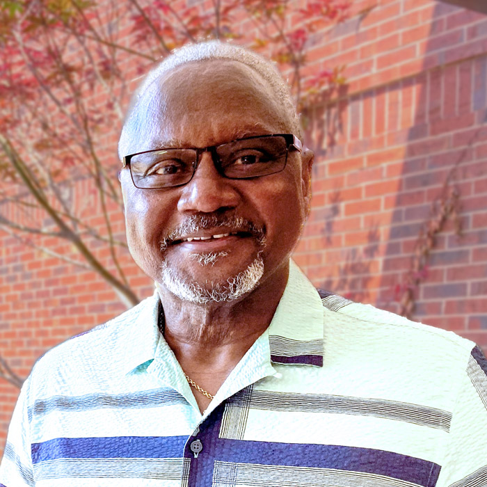 A smiling man wearing glasses stands outside in front of a red-brick wall.