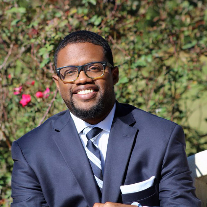 A smiling man in a blue suit stands in front of shrubbery.