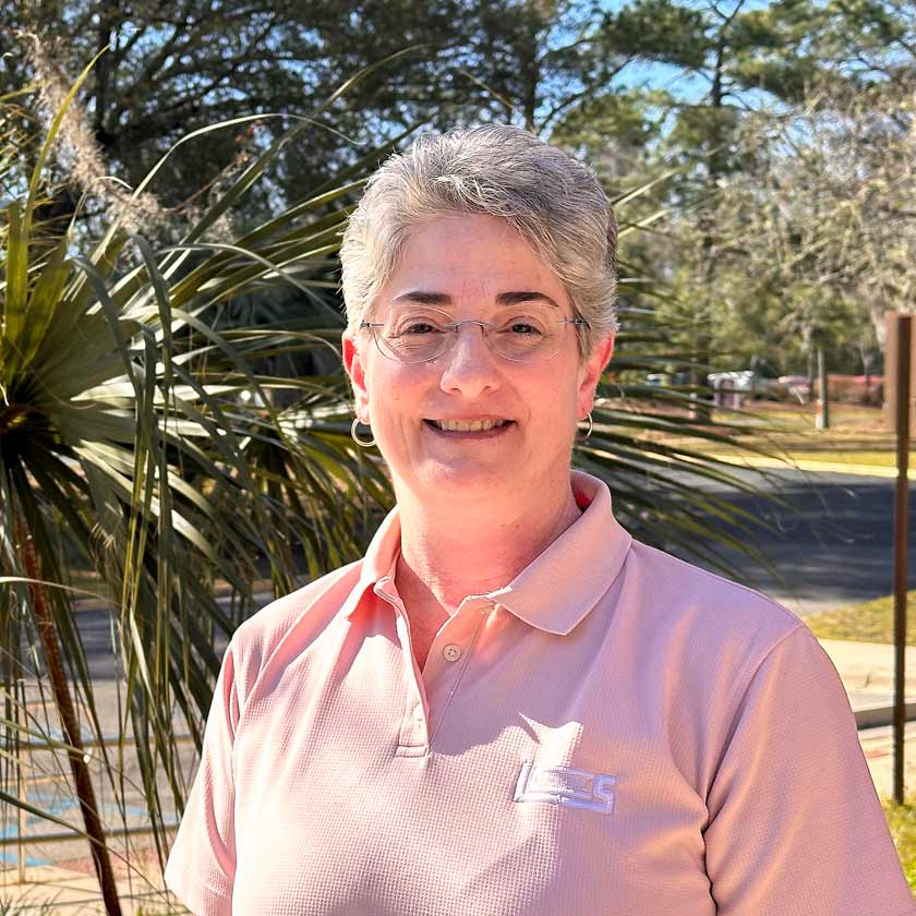 A smiling woman in a pink shirt stands in front of shrubbery.