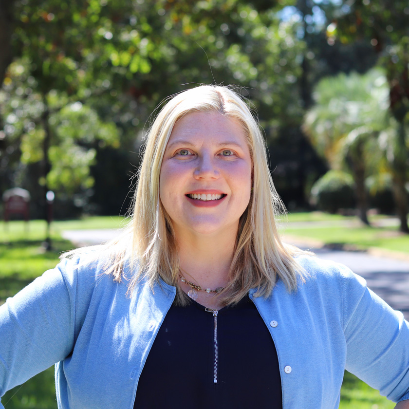 A smiling woman stands otside in front of a tree.