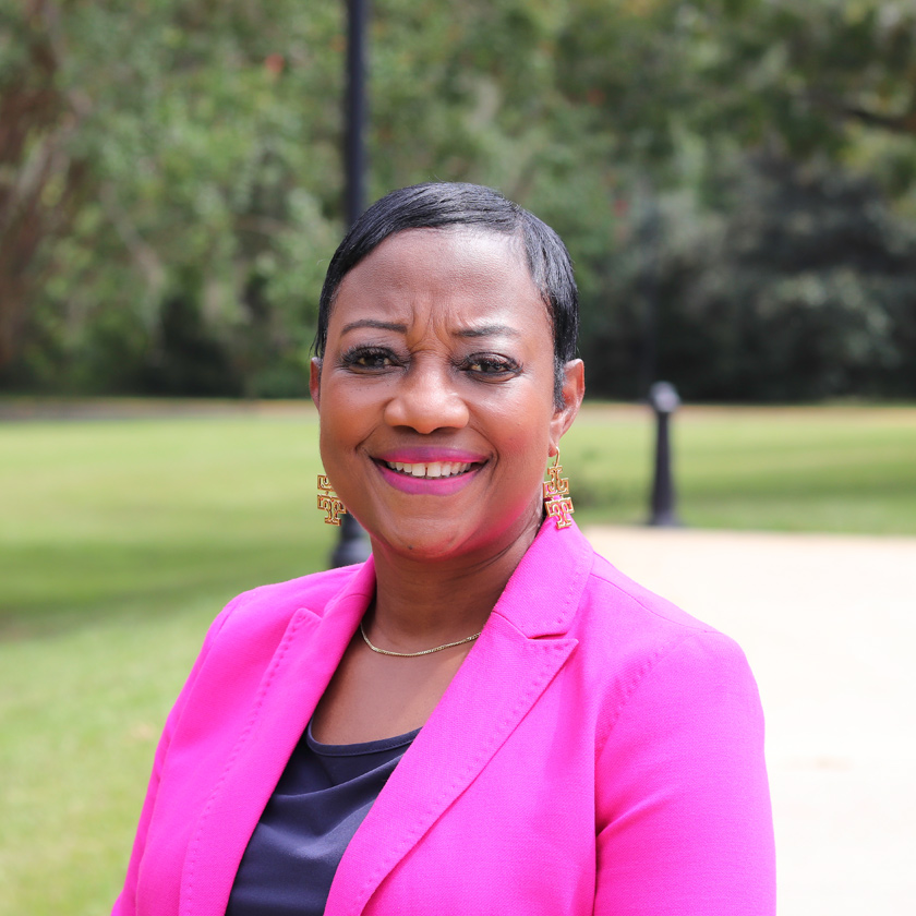 A smiling woman dressed in pink stands in front of shrubbery