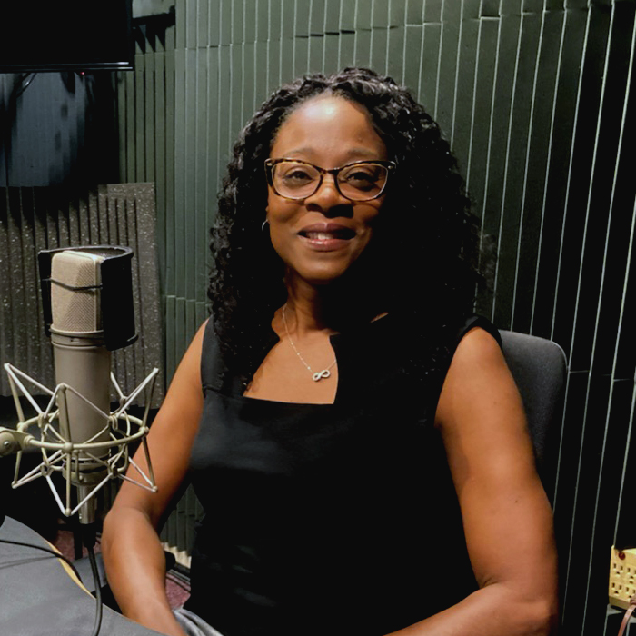 A smiling woman dressed in black sits at a table in front of a microphone. 