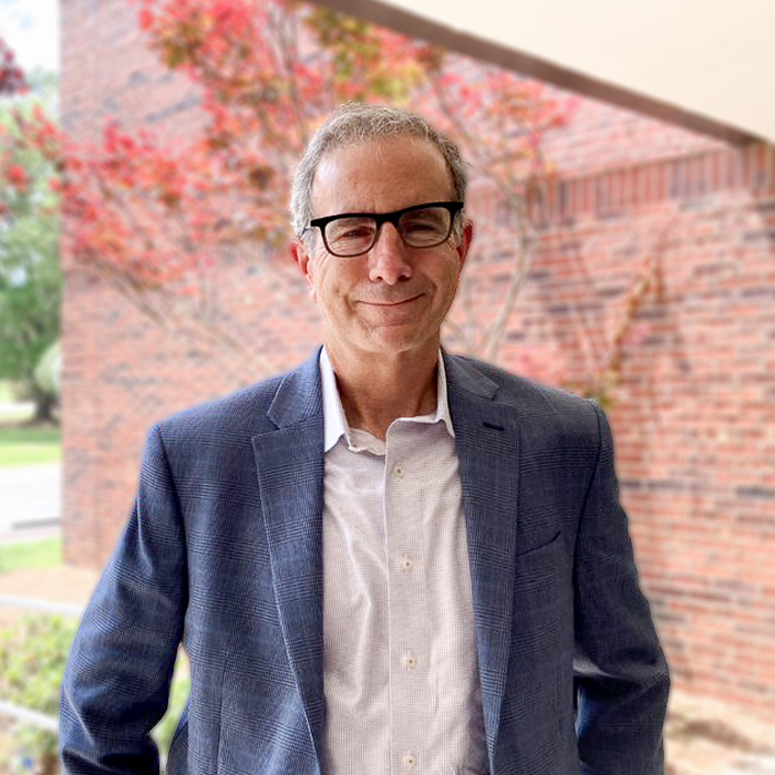 A smiling man in a blue suit stands outside in front of a red brick wall