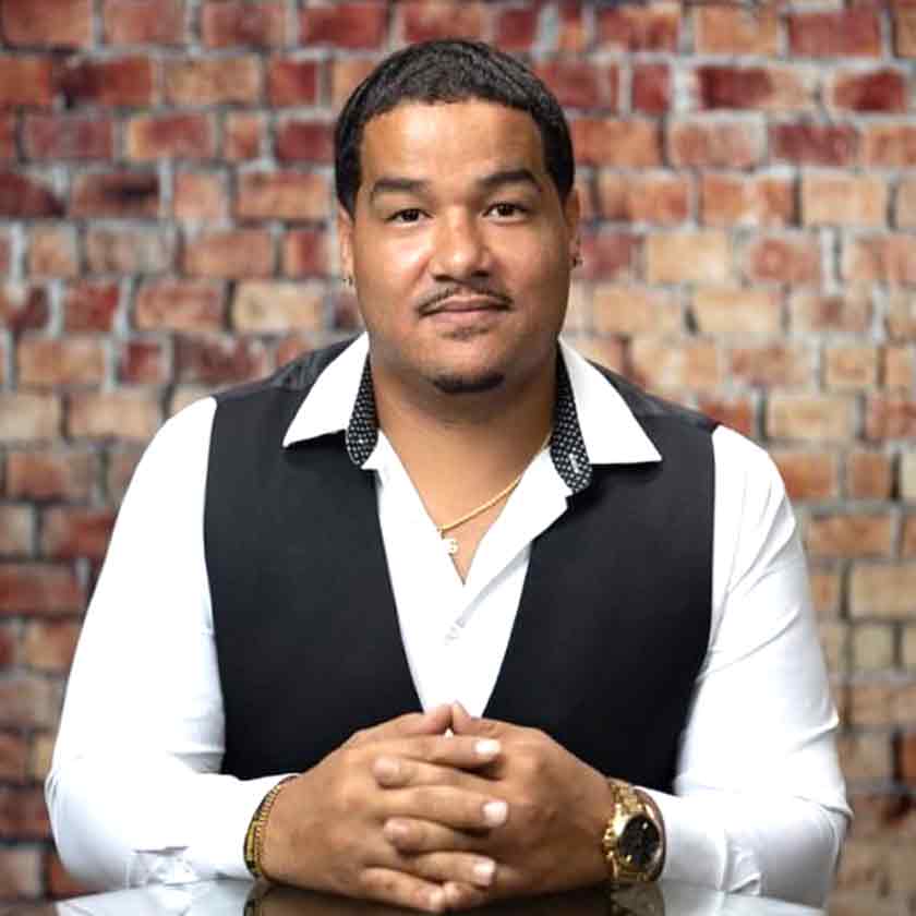 A smiling man sits with hands folded in front of a red brick wall.