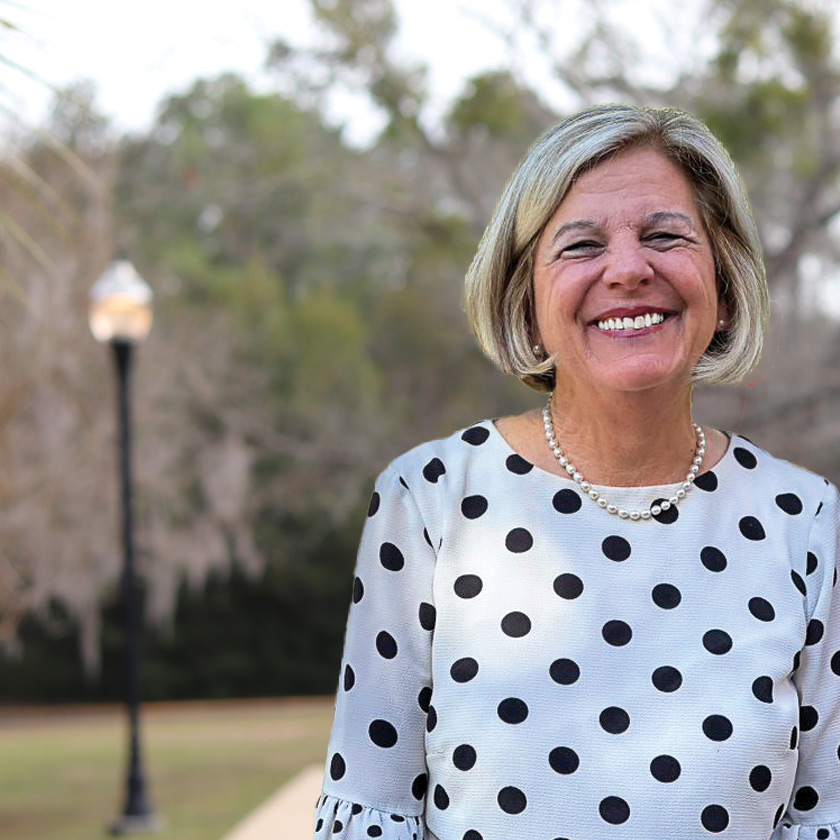 A smiling woman in a white and black top stands in front of shrubbery.