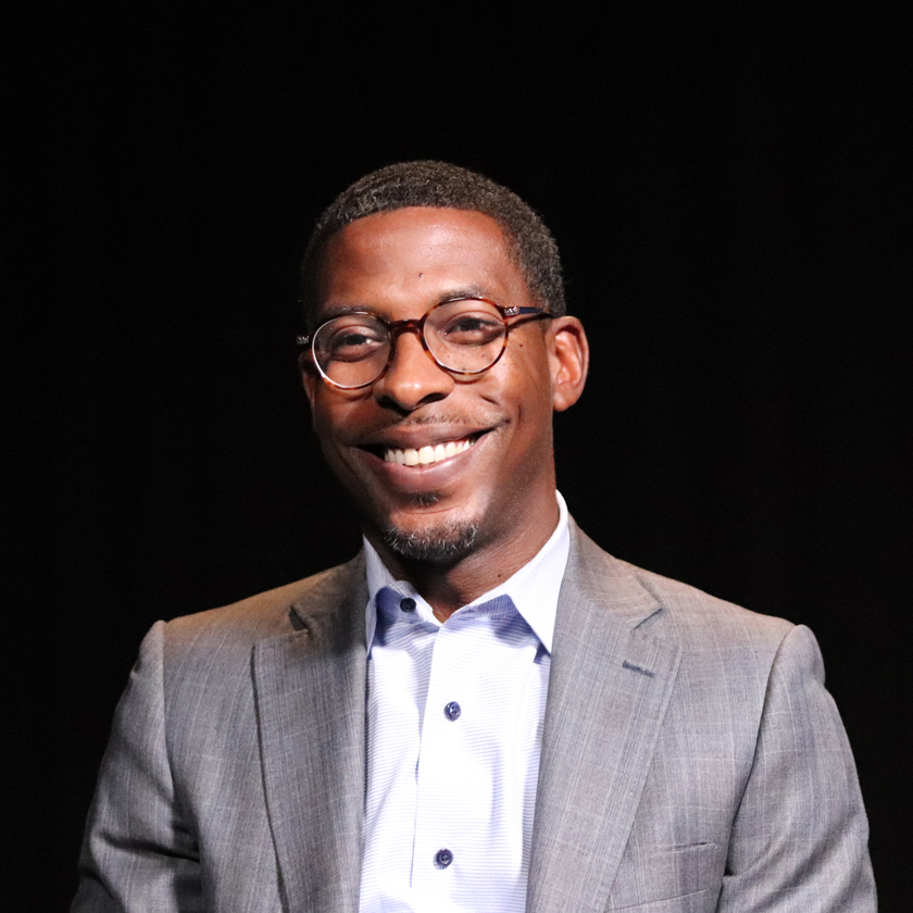 A smiling man in a grey suit sits in front of  a dark background.