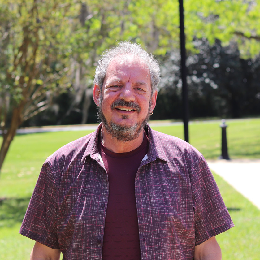 A smiling man stands outside in front of shrubbery.