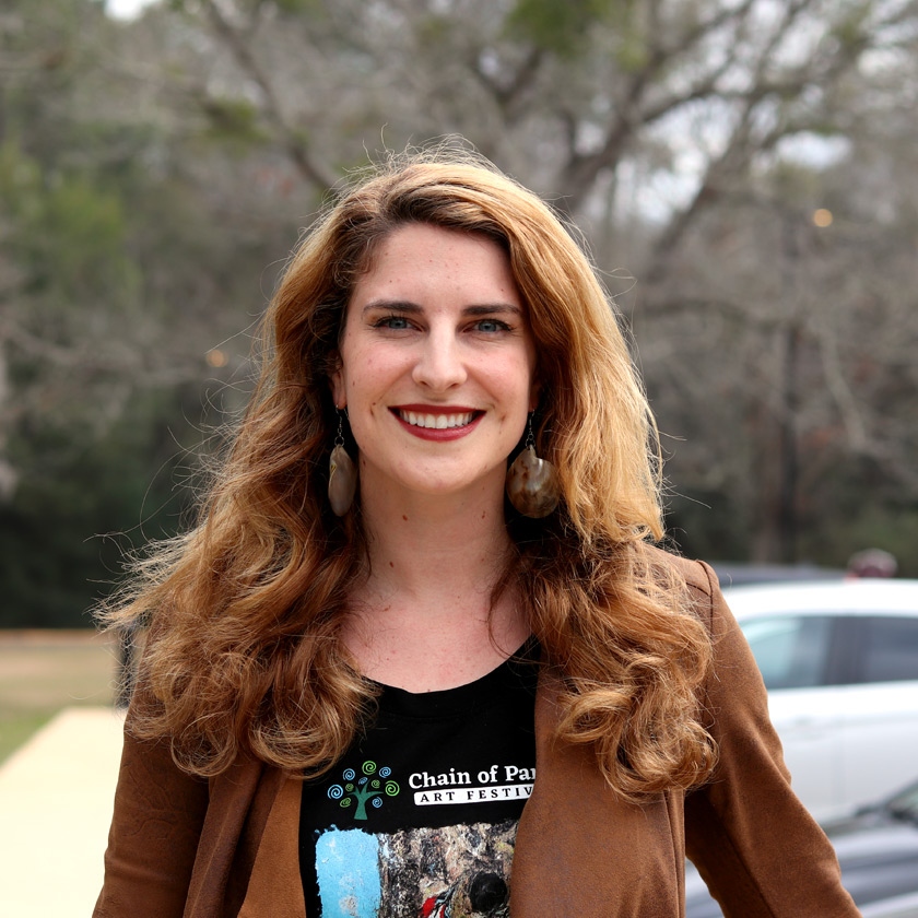 A smiling woman stands in front of shrubbery.