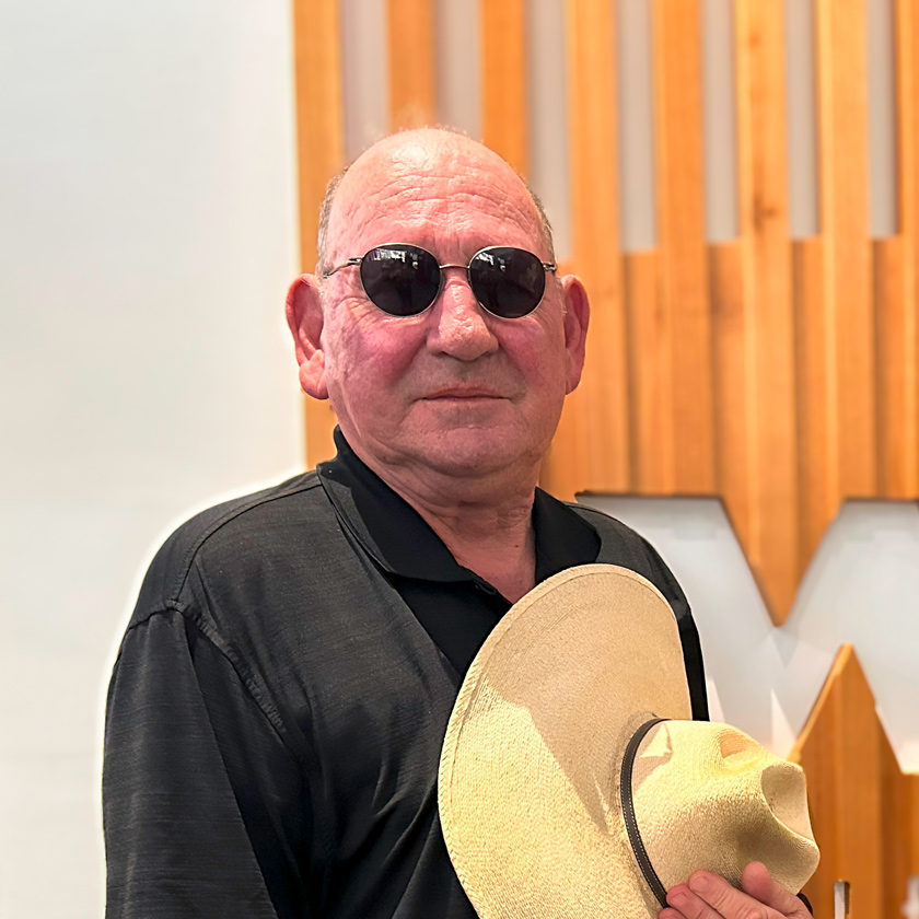 A smiling man in shades stands in front of a white wall and a wooden structure.