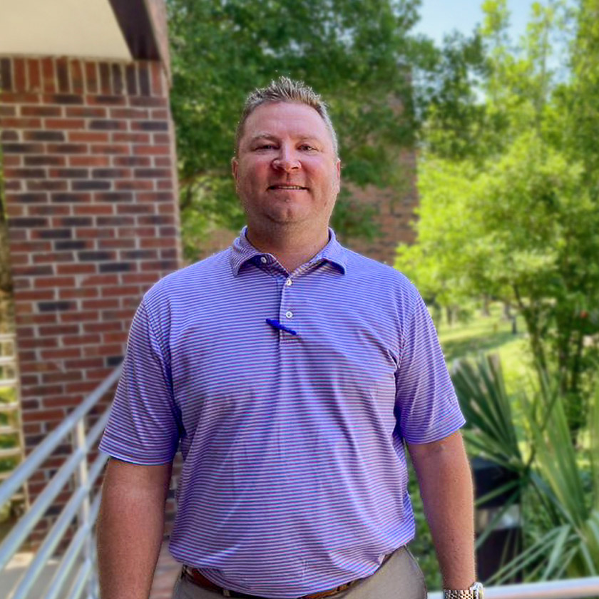 A man in a purple shirt stands outside smiling.