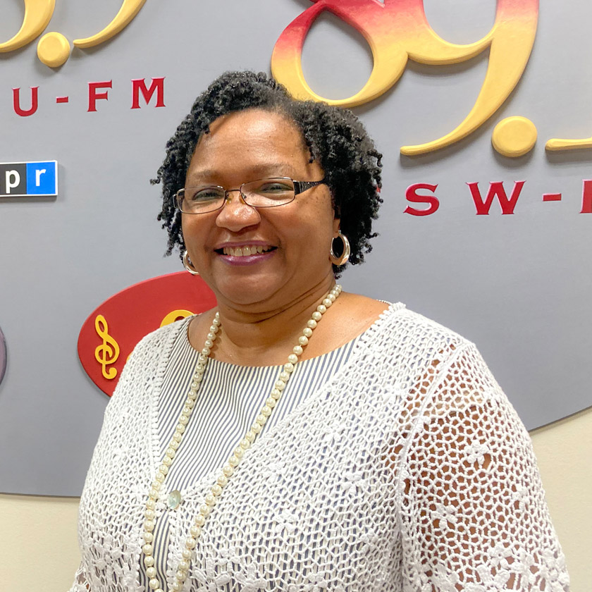 A smiling woman in a white blouse stands in front of a grey, garnet and gold mural.