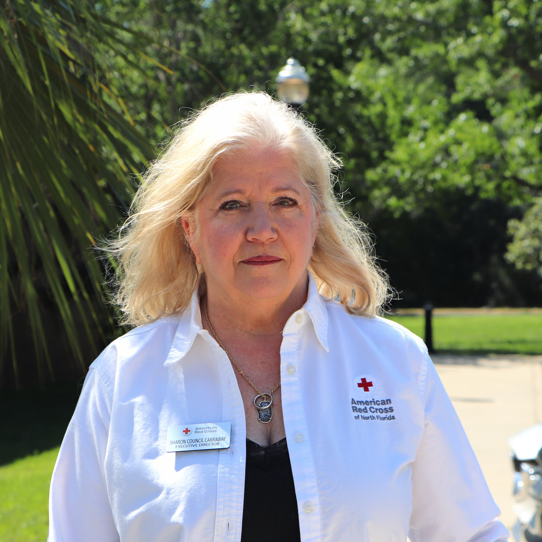 A woman in a white shirt stands outside in front of shrubbery.