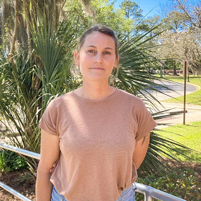 A smiling woman stands in front of green shrubbery. 