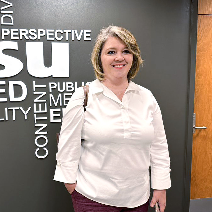 A smiling woman dressed in a white shirt stands in front of a gray wall.