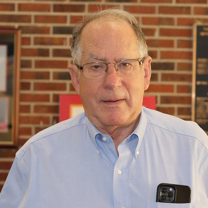 Bespectacled man in light button-up shirt stand in front of a red brick wall.