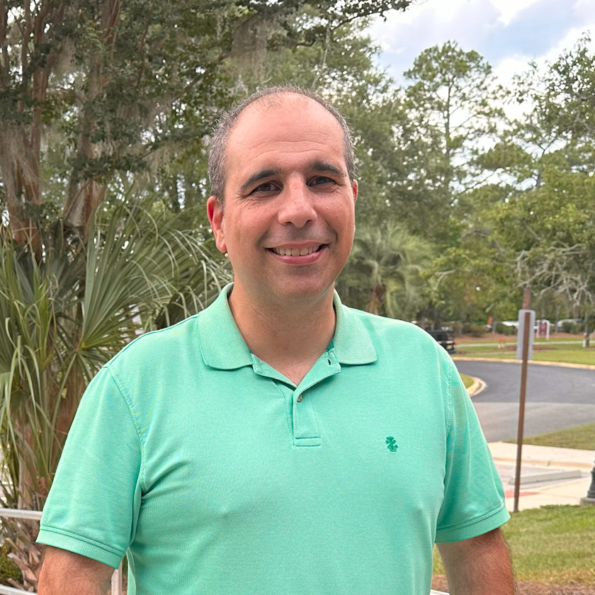 A smiling man stands in front of shrubbery.