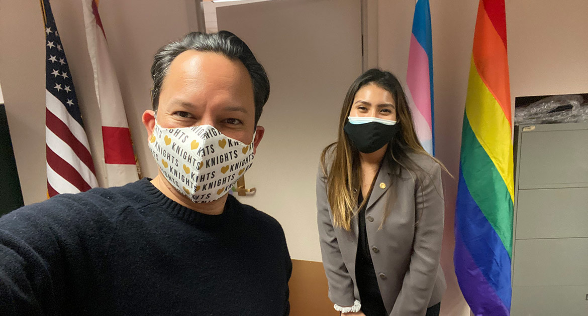 man and woman in mask standing next to flags (gay pride) taking selfie