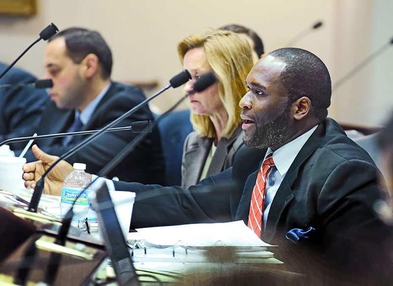 government looking man in suit looking left, sitting behind microphone