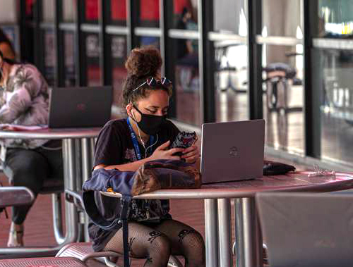 young woman sitting at outside table working at laptop, wearing mask